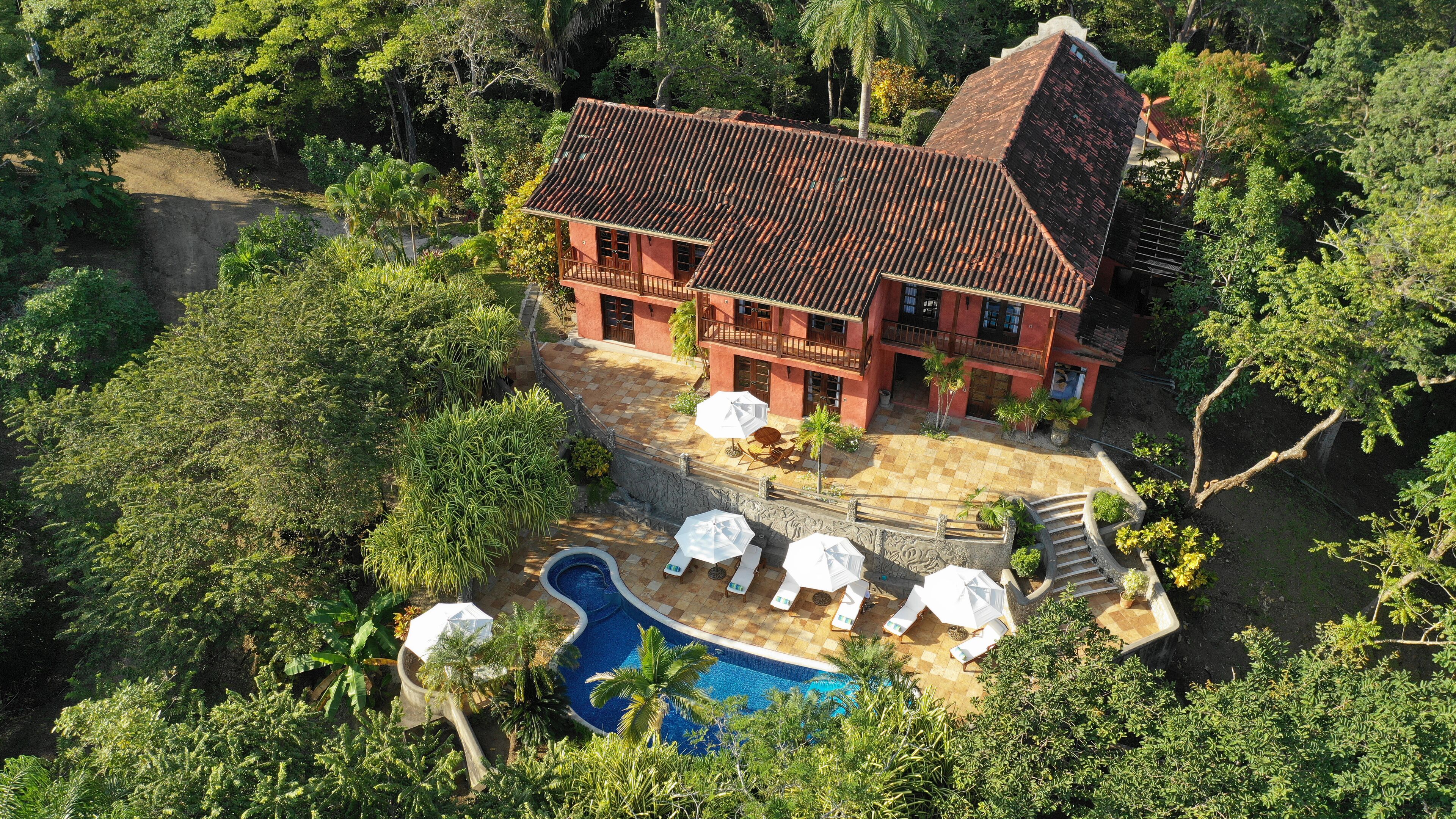 Una villa grande con techo rojo, piscina y zona de estar exterior rodeada de exuberante vegetación.
