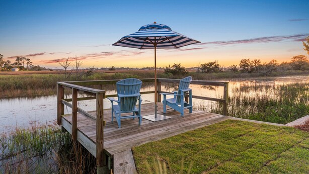 Un pontile in legno con due poltrone Adirondack e un ombrellone a righe, con vista su un'acqua calma.