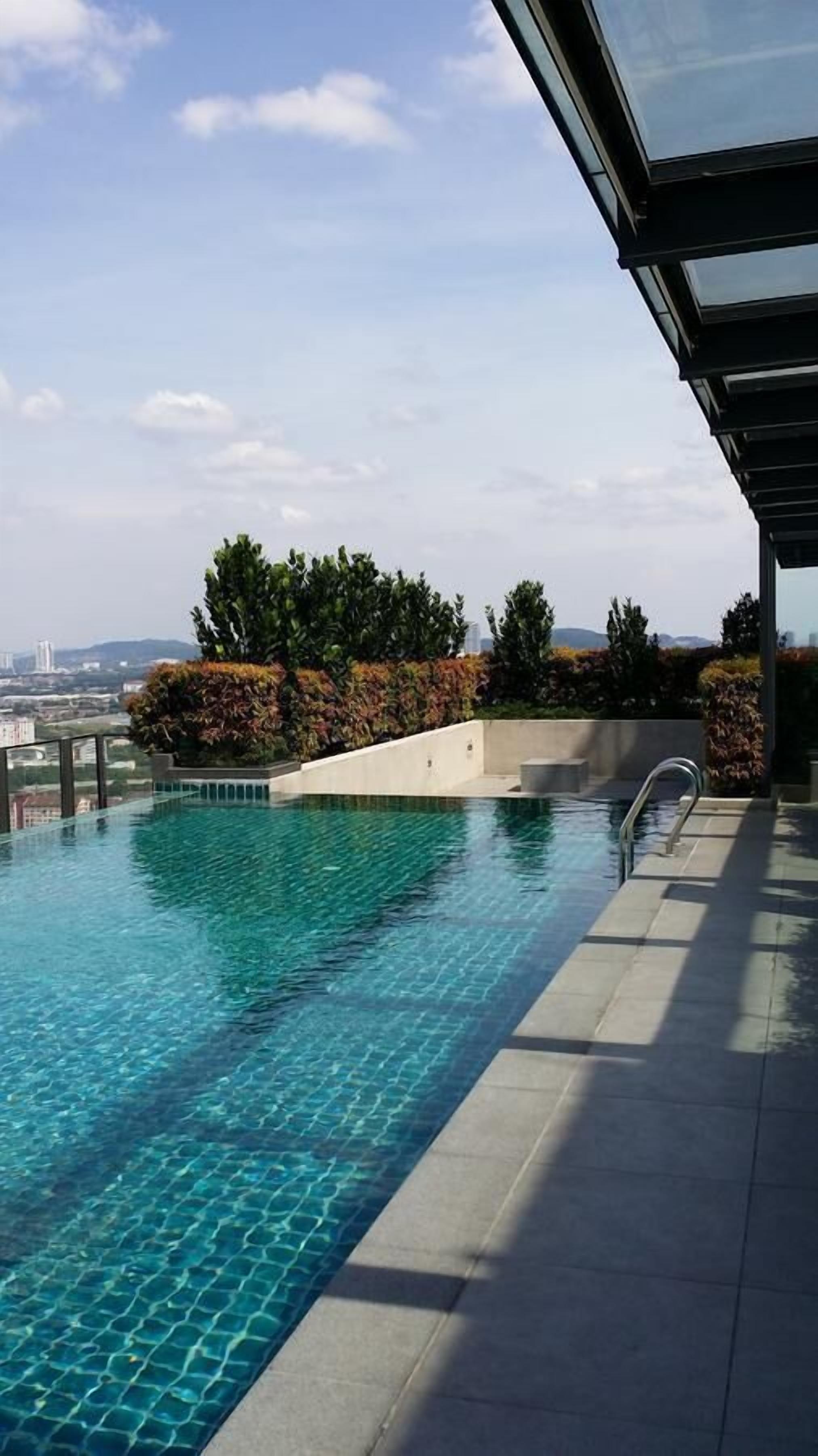 An infinity pool with clear blue water, surrounded by a modern architectural structure with a glass roof, and a view of distant buildings and hills.