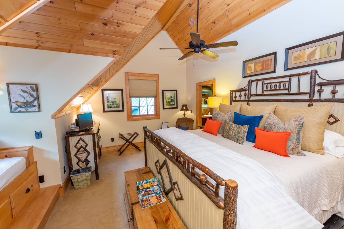 A bedroom with a large bed, wooden headboard, and a ceiling fan. There are framed pictures on the wall, a desk with a lamp, and a window with blinds.
