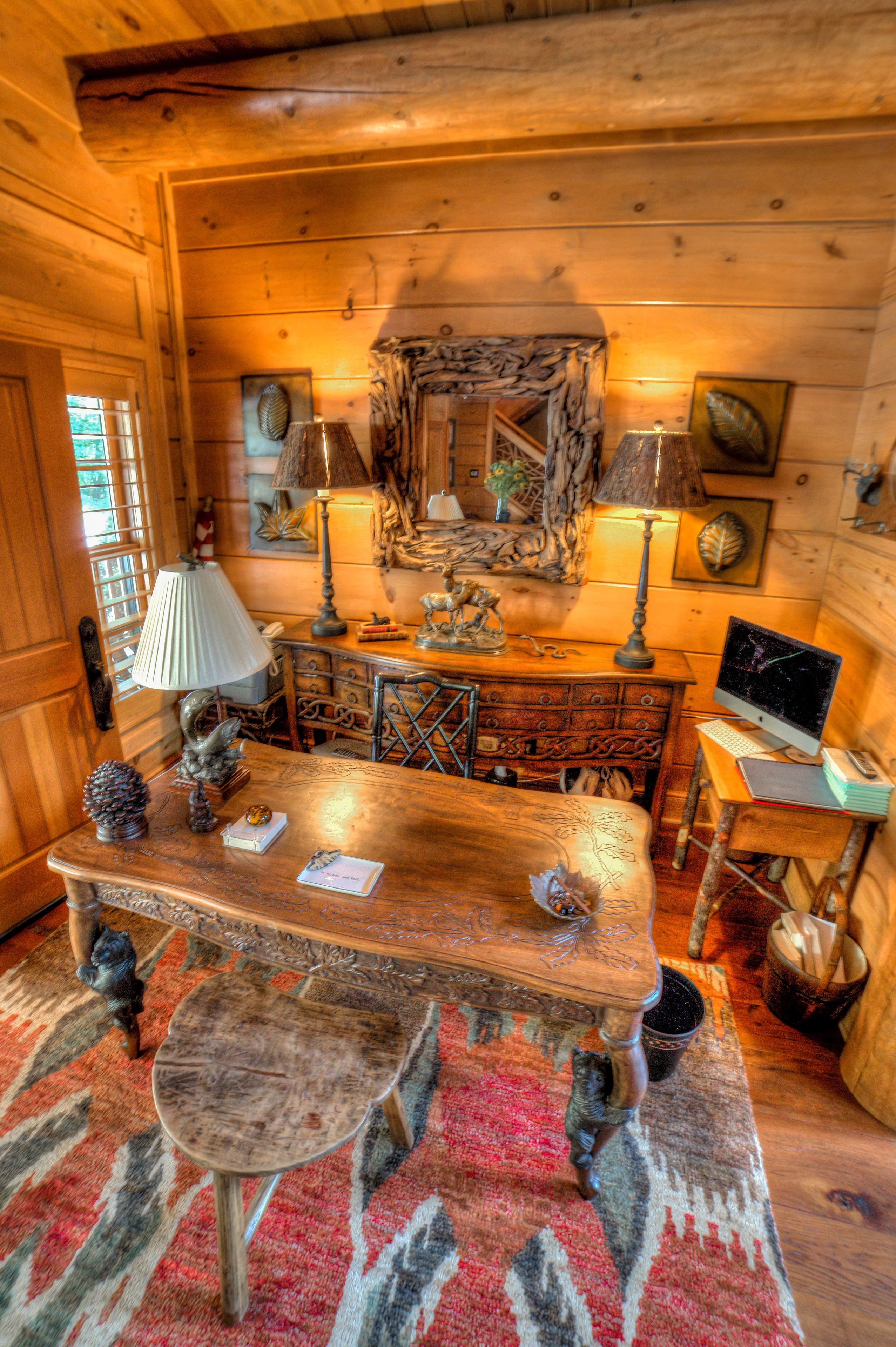 A rustic wooden room with a wooden table, a chair, a lamp, a computer, and a decorative mirror.