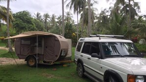 A white SUV parked next to a beige tent in a grassy area with palm trees in the background. - IDestinos Tayrona mobile home  (La Poza)