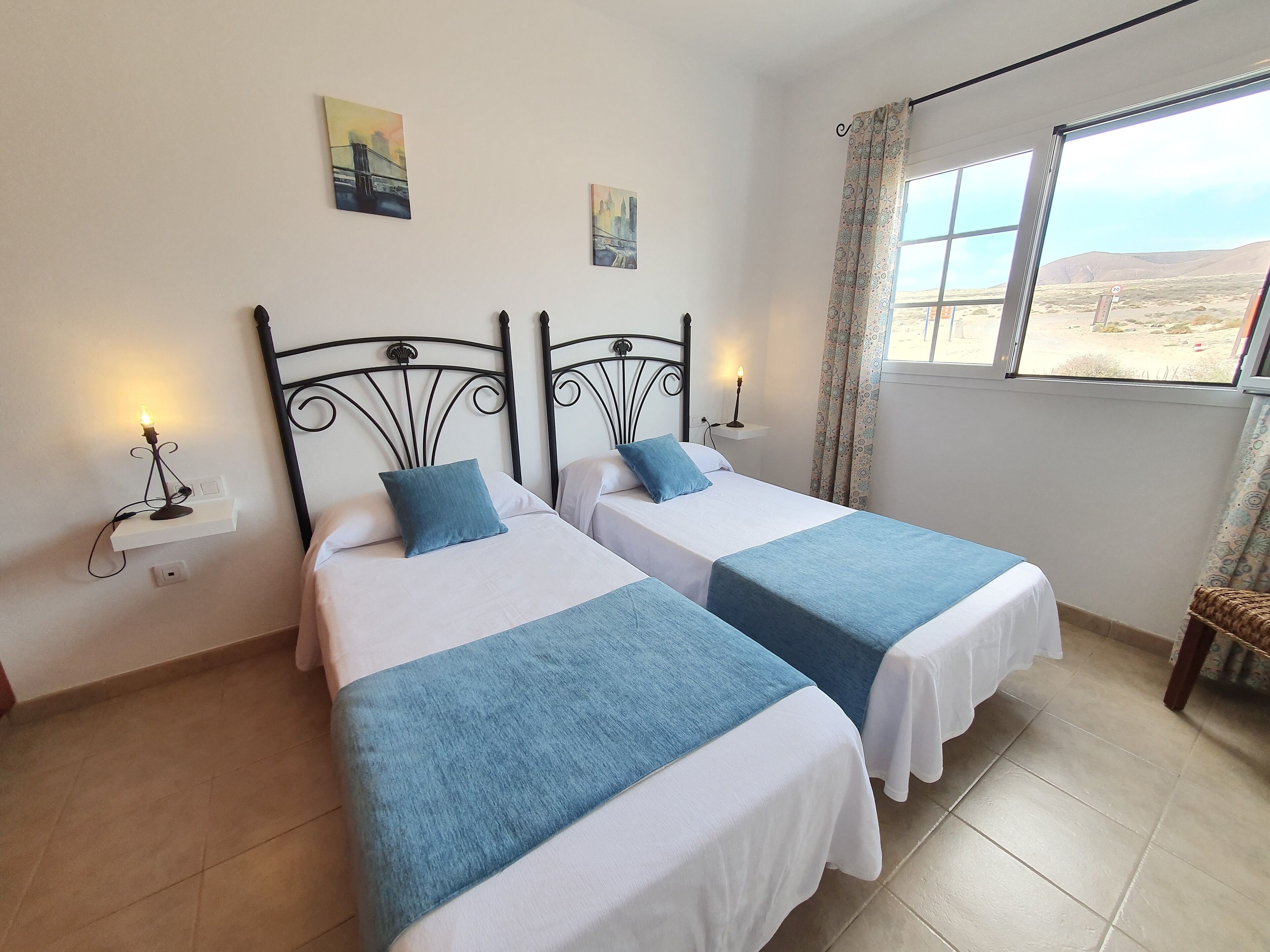 A hotel room with two beds, each with a blue bedspread and white linens. There are wall-mounted nightstands with lamps, and two paintings above the beds. A window with patterned curtains provides a view of the desert landscape.