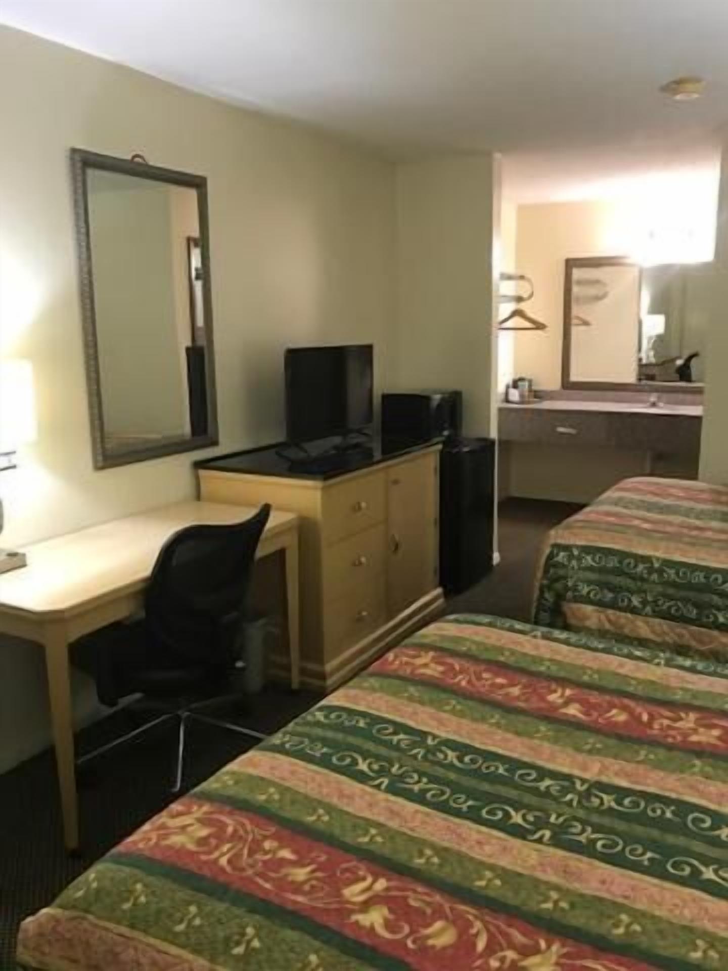 A hotel room with a desk, a television, a bed with patterned bedding, a dresser, and a mirror.
