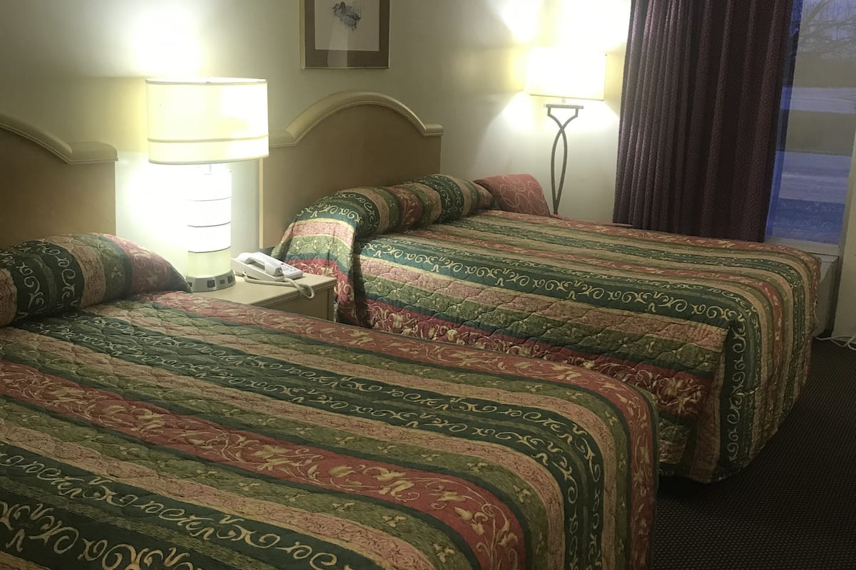A hotel room with two beds, a nightstand, a lamp, and a window with curtains.