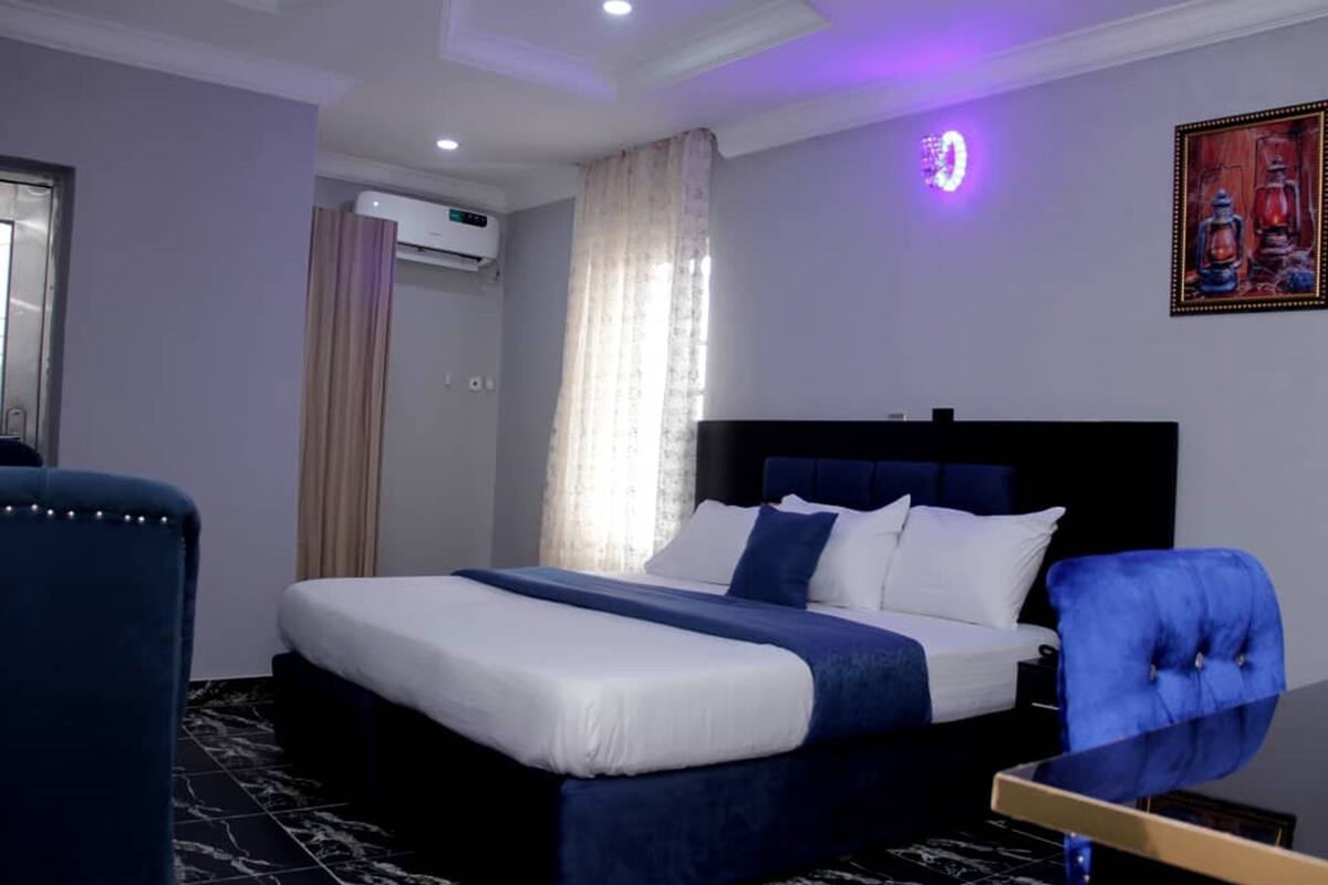 Une chambre d’hôtel comprenant un lit, une chaise, une fenêtre avec des rideaux, un appareil de climatisation et un luminaire fixé au mur.