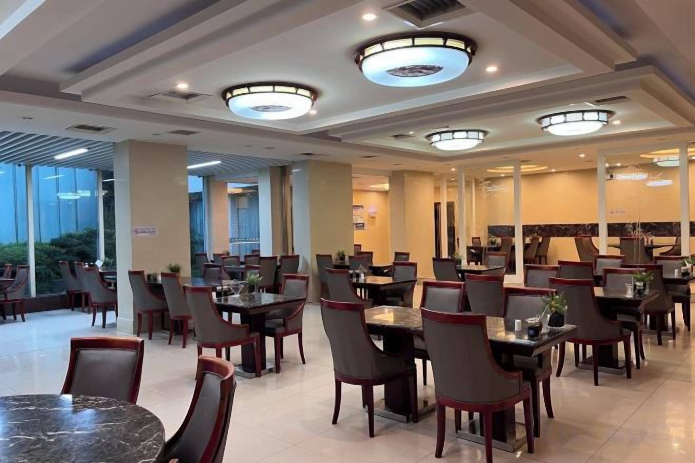 A hotel dining area with multiple tables and chairs, large windows, and ceiling lights.