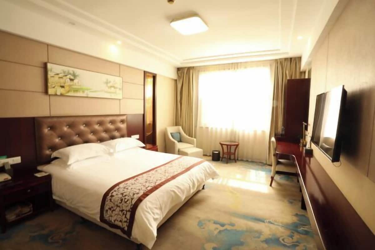A hotel room with a large bed, a desk, a chair, a small table, a television, and a window with curtains.