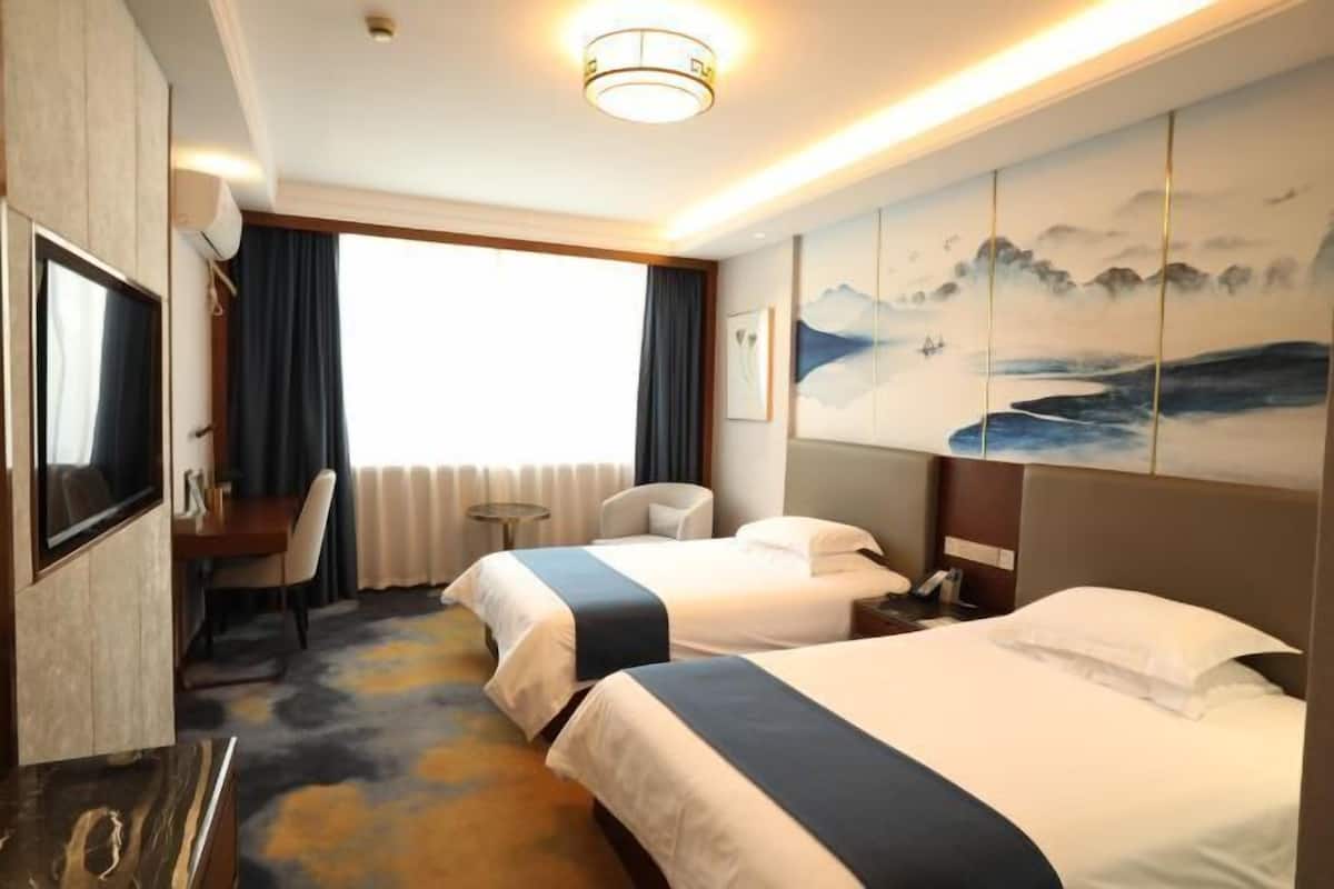 A hotel room with two beds, a desk, a chair, a TV, and artwork on the wall.