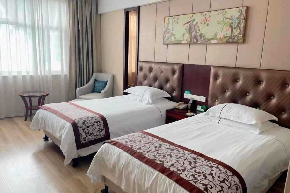A hotel room with two beds, a chair, a small table, and a painting on the wall.