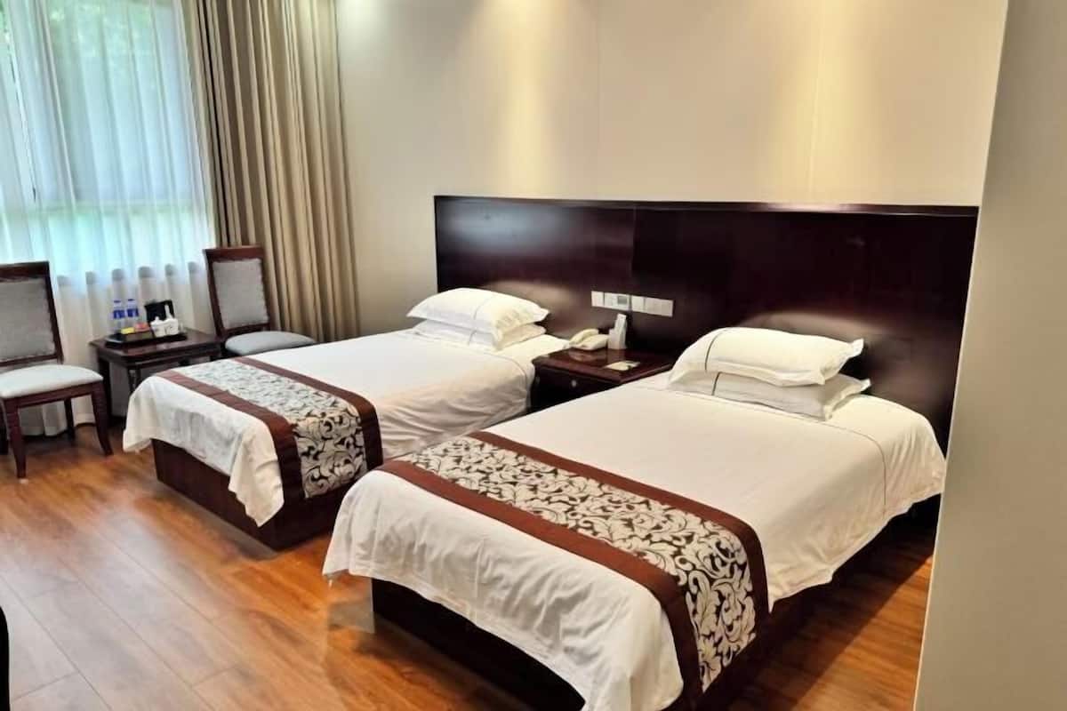 A hotel room with two beds, wooden flooring, a large window with curtains, and a bedside table with a lamp.