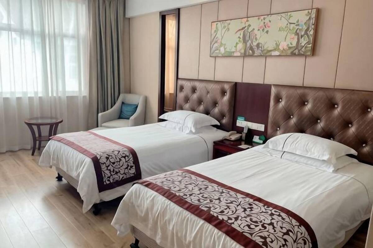 A hotel room with two beds, a chair, a small table, and a painting on the wall.