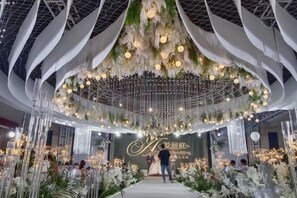 An indoor event space with a central stage, white drapery, hanging floral arrangements, and guests seated at tables. - Huaian Hotel (Huai'an)
