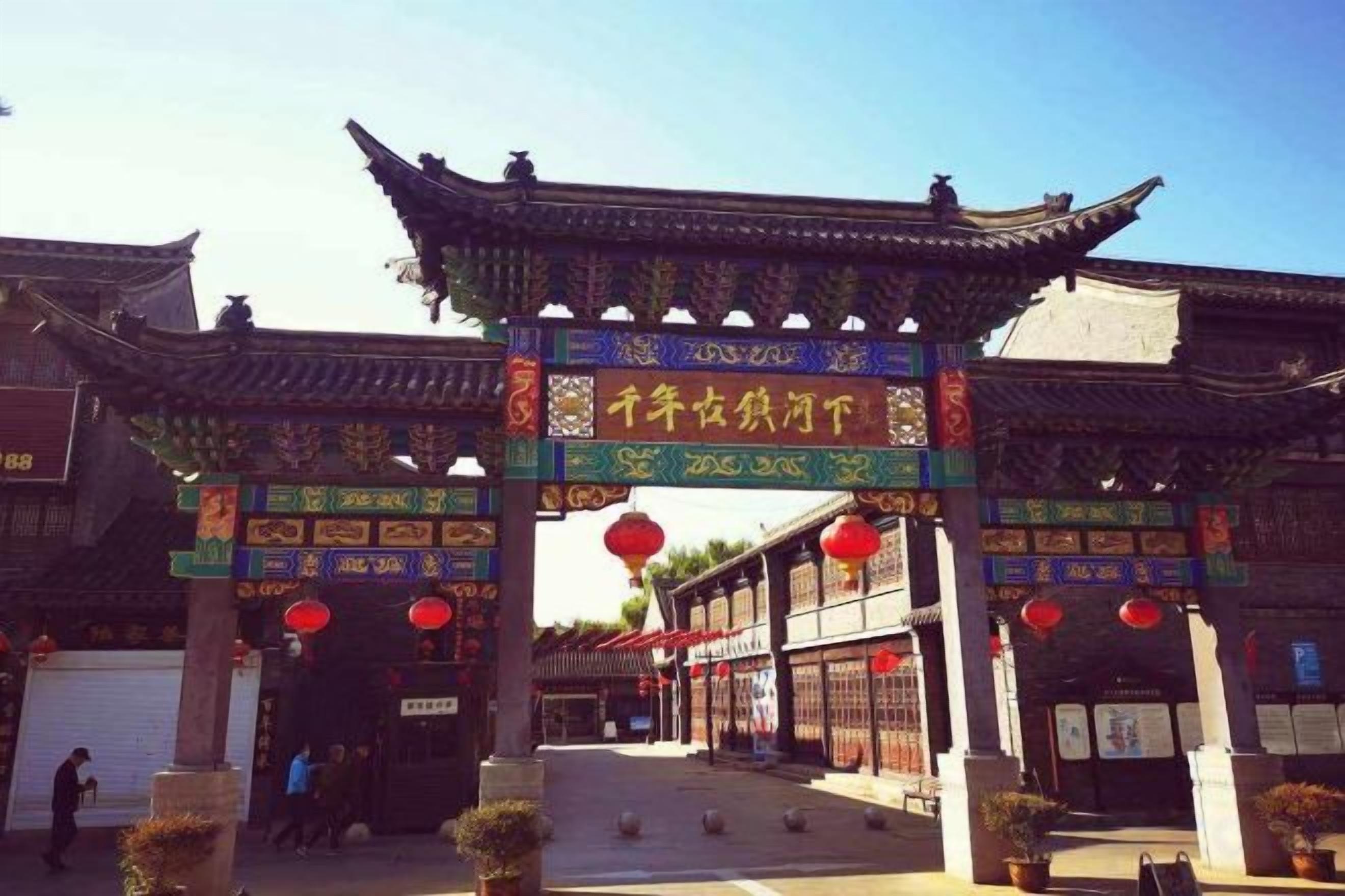A traditional Chinese gate with intricate designs and characters, leading to a courtyard with potted plants and a clear sky.