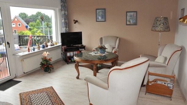Uma sala de estar com televisão, uma mesa de centro redonda, duas poltronas, um sofá e uma janela com vista para uma casa e árvores.