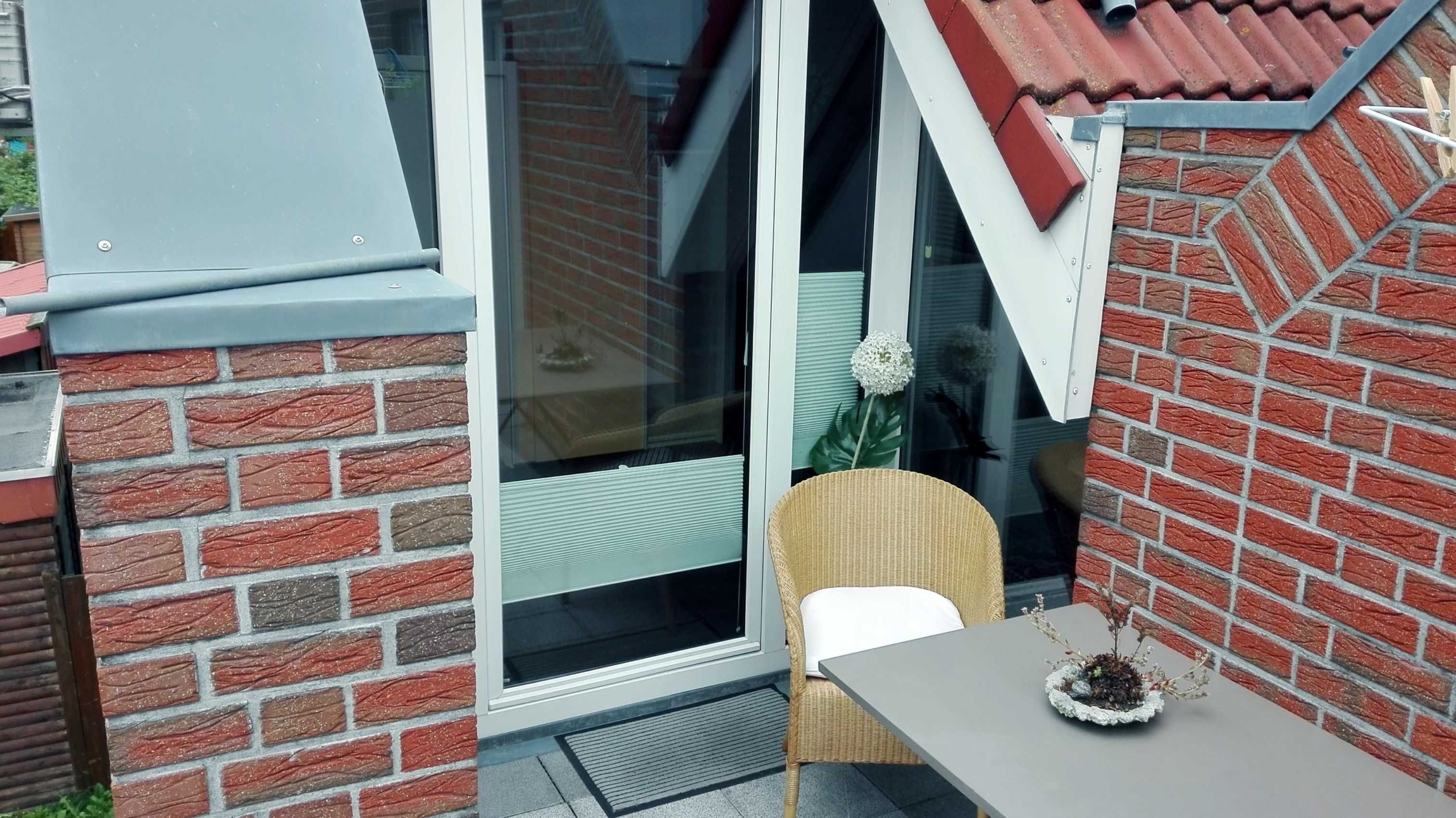 Ein kleiner Sitzbereich im Freien mit einem Korbstuhl und einem Tisch, einer Ziegelwand und einer Glastür, die auf eine überdachte Terrasse führt.
