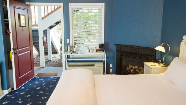A bedroom with a bed, a nightstand, a lamp, a fireplace, a window with blinds, a door, and a staircase leading to an outdoor area. - The Inn On First (Napa)