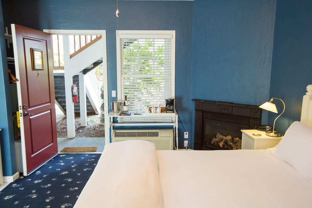 A bedroom with a bed, a nightstand, a lamp, a fireplace, a window with blinds, a door, and a staircase leading to an outdoor area.