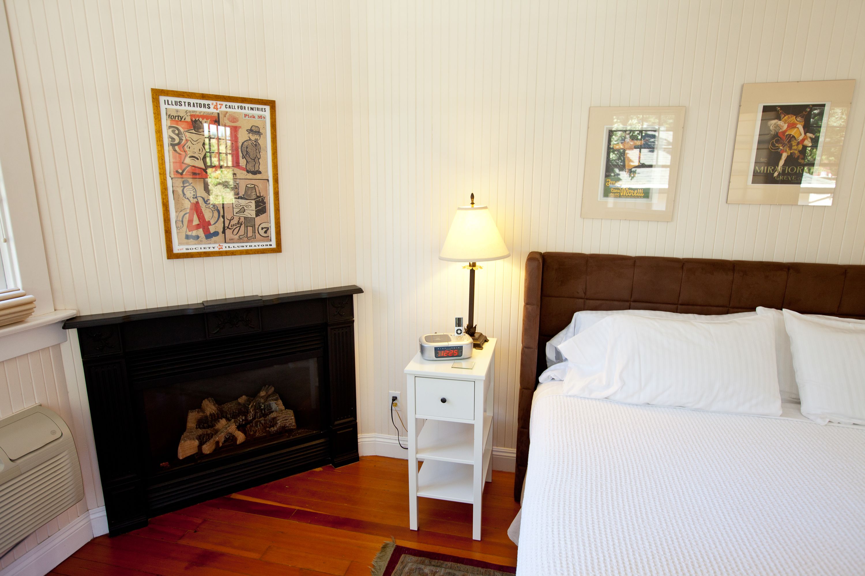 A bedroom with a fireplace, a bedside table with a lamp, a bed with white bedding, and framed pictures on the wall.