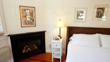 A bedroom with a fireplace, a bedside table with a lamp, a bed with white bedding, and framed pictures on the wall.