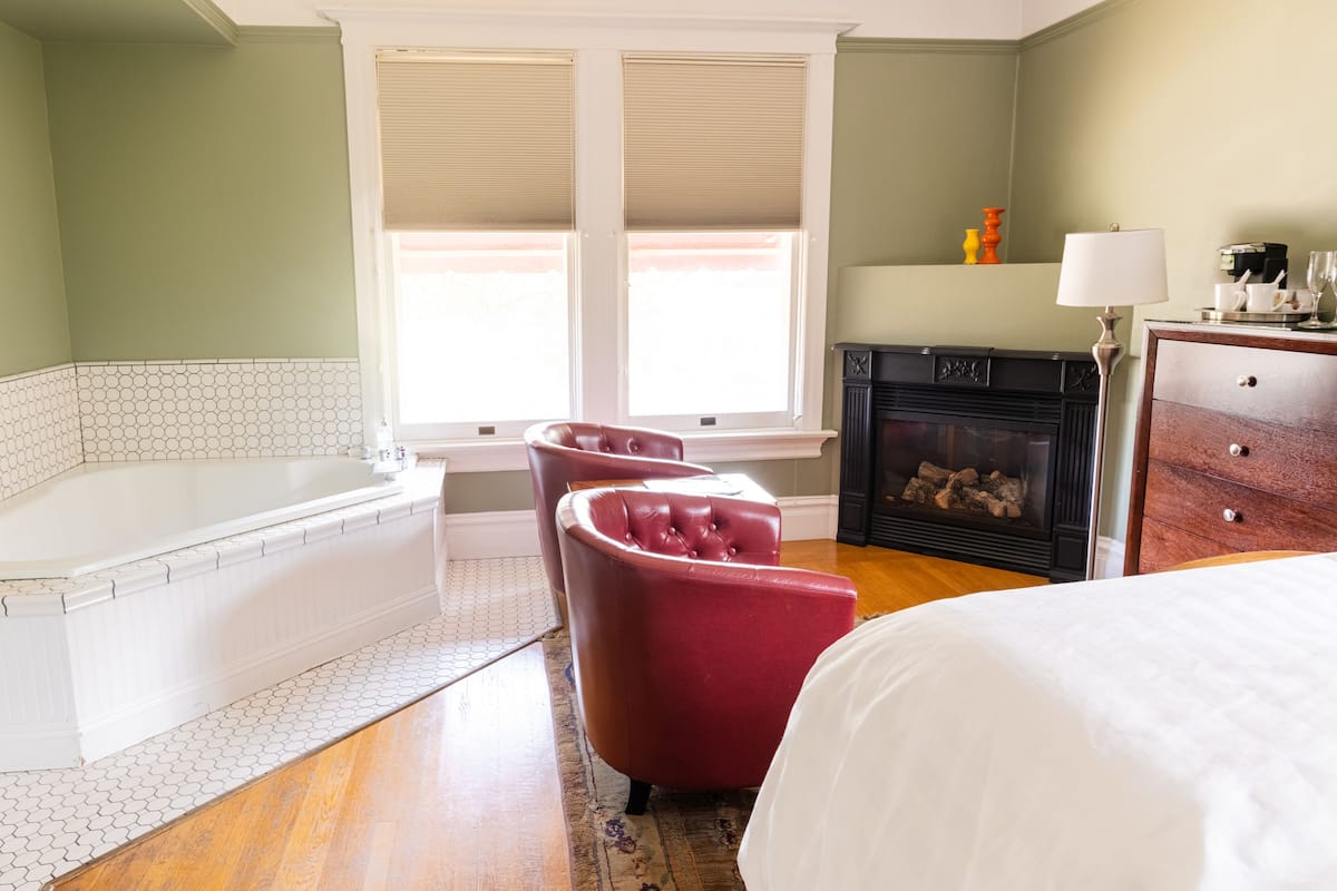 A bedroom with a large bed, a red armchair, a white bathtub, and a fireplace.