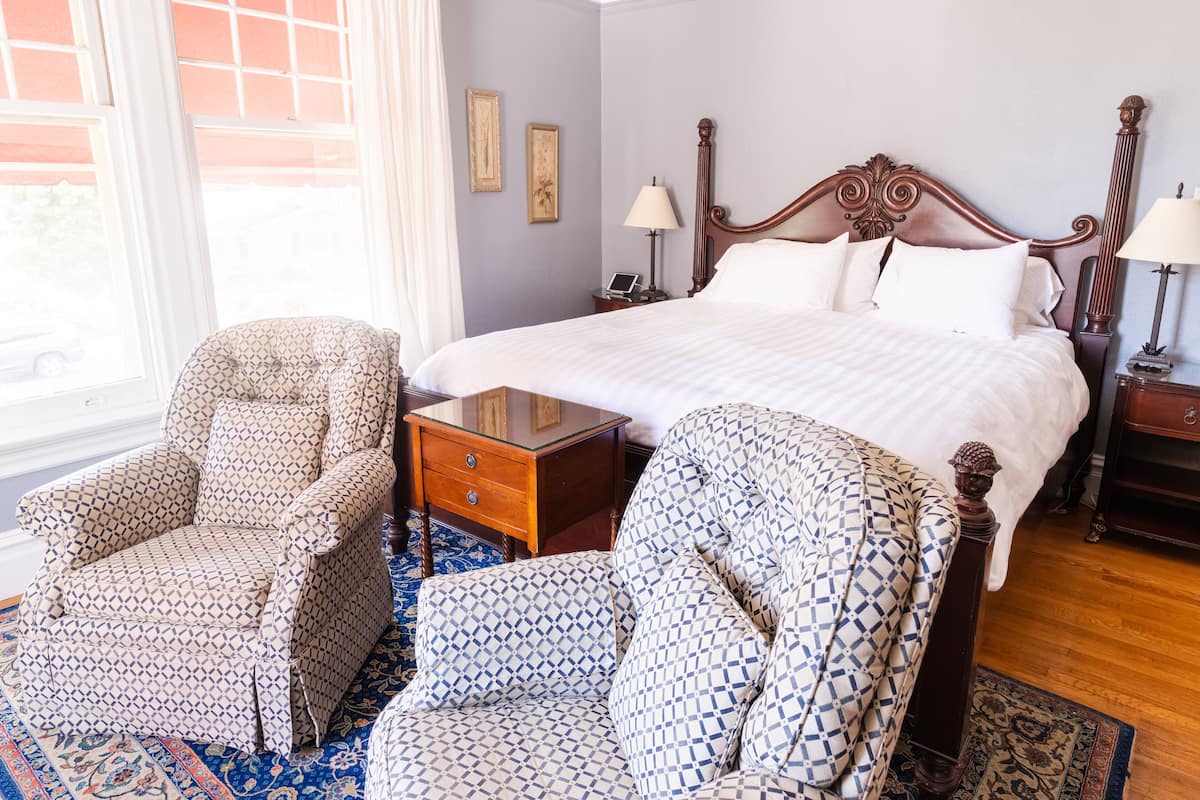A hotel room with a large bed, two armchairs, a wooden nightstand, and a window with curtains.