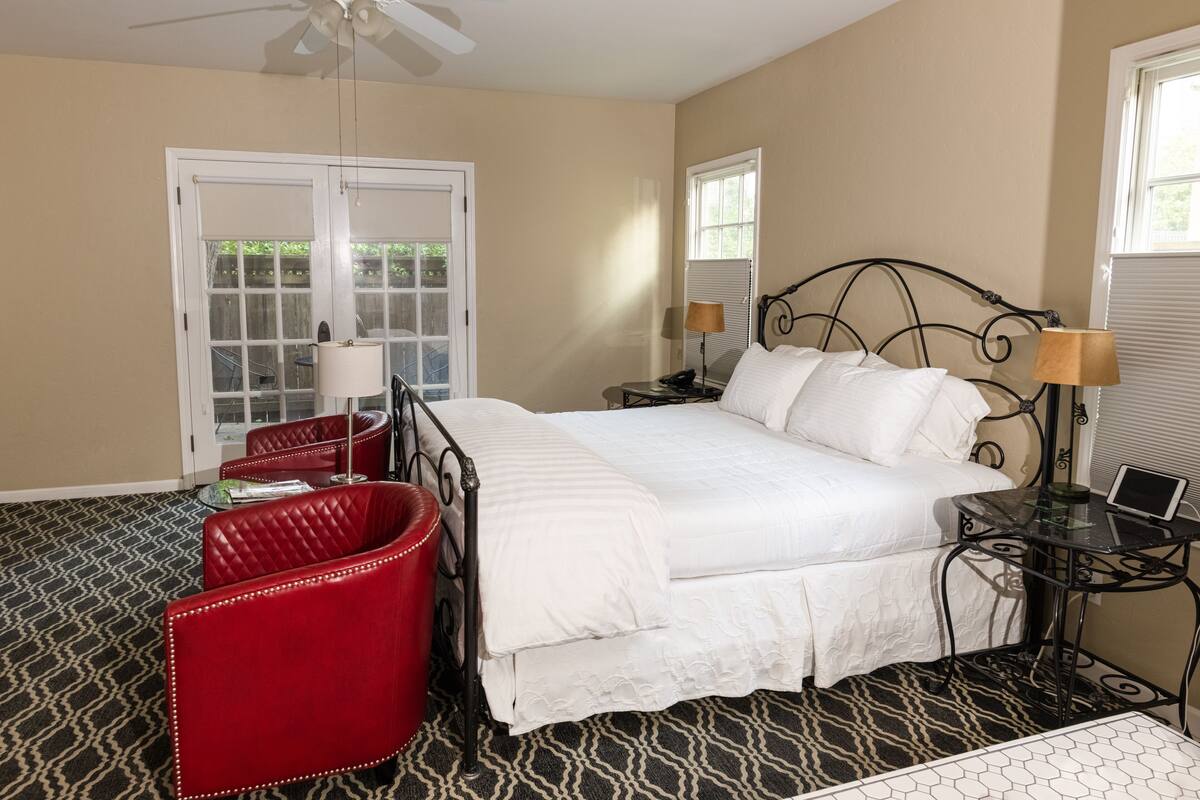 A hotel room with a bed, a red chair, a desk with a lamp, and a ceiling fan.
