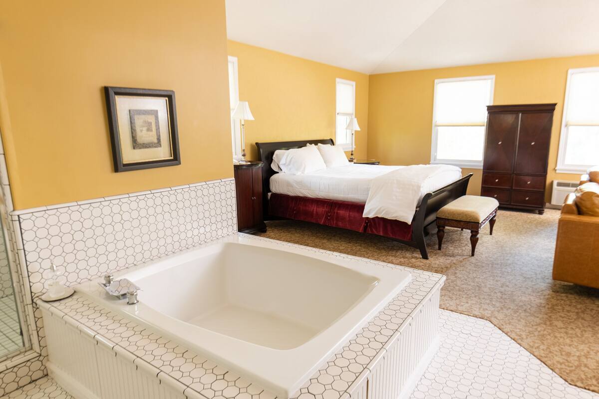 A bedroom with a large white bathtub, a bed with white linens, a wooden nightstand, and a wardrobe.