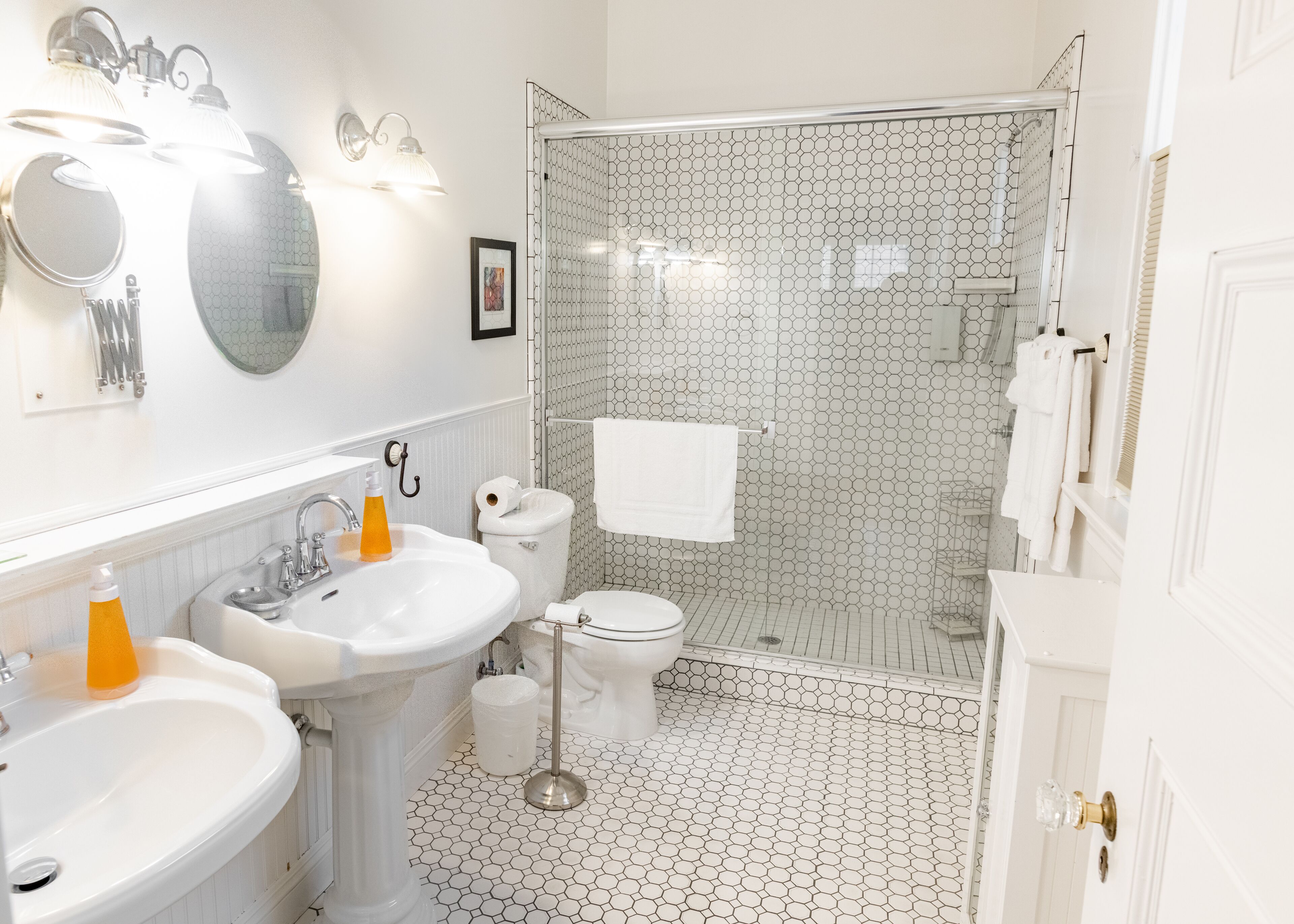 Une salle de bain avec un lavabo, des toilettes et une douche, le tout de couleur blanche. Un miroir est placé au-dessus du lavabo et une serviette est accrochée à la porte de la douche.