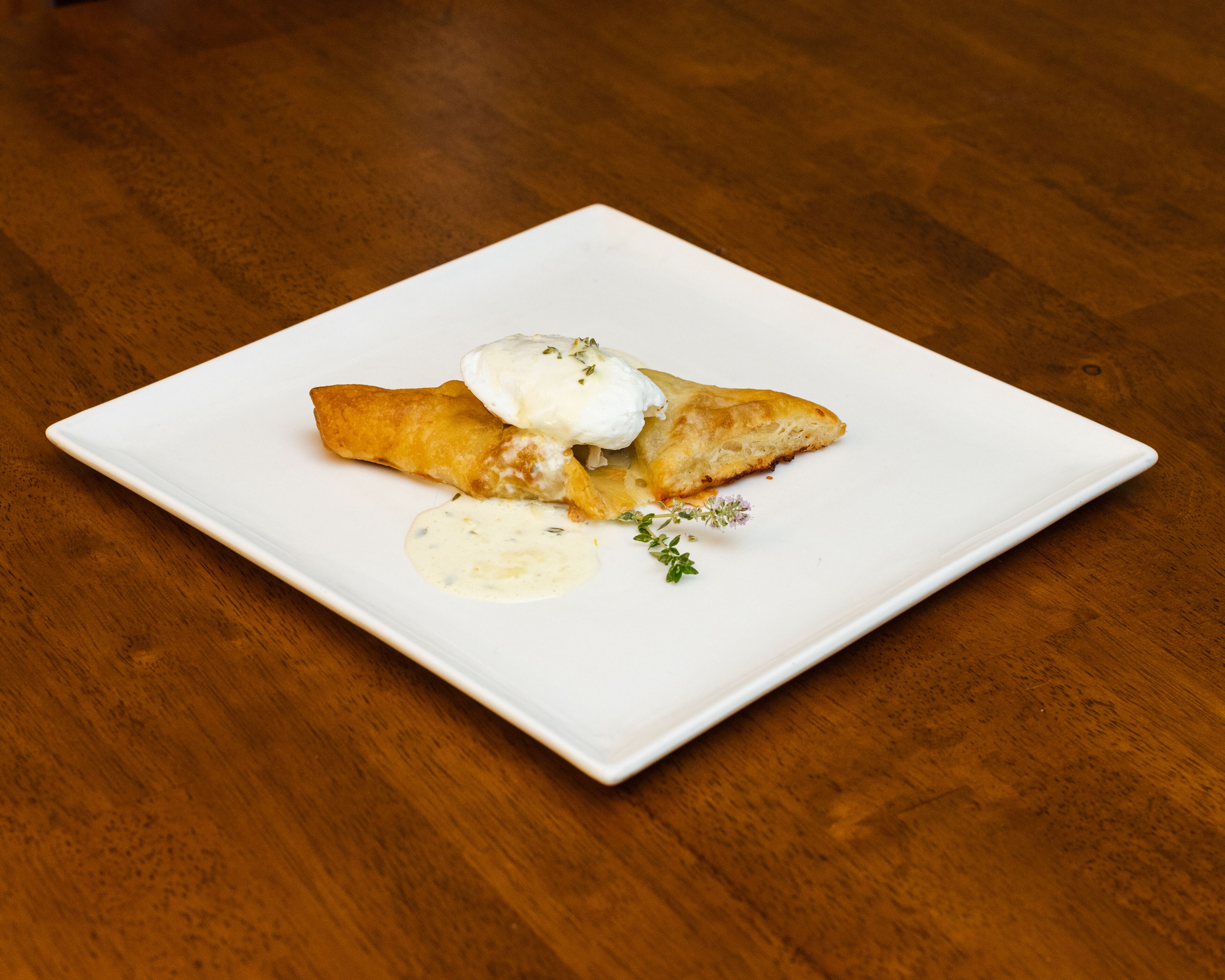 Un plato cuadrado blanco con un trozo de comida frita, posiblemente un croissant, cubierto con salsa blanca y adornado con hierbas.