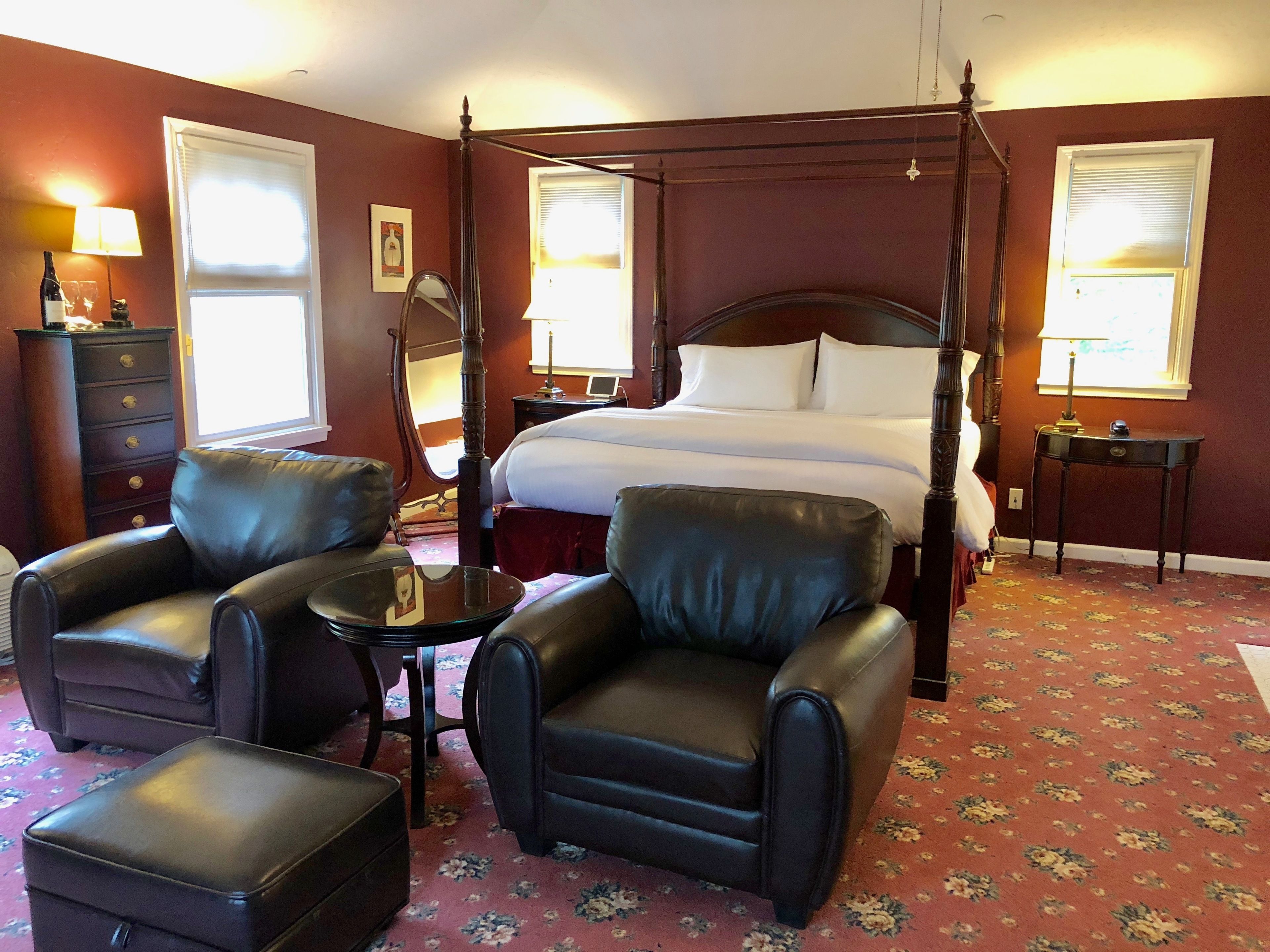 A four-poster bed with white linens, a dark wooden headboard, and a floral carpet. There are two leather armchairs and a small table with a lamp.