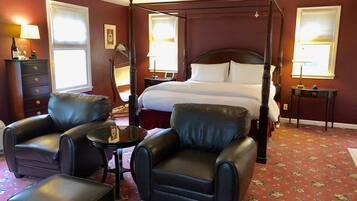 A four-poster bed with white linens, a dark wooden headboard, and a floral carpet. There are two leather armchairs and a small table with a lamp.
