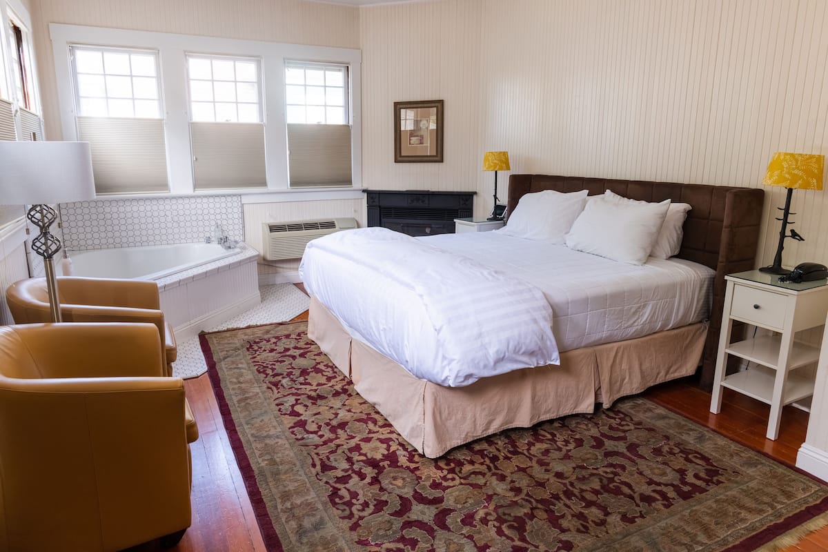 A hotel room with a bed, a sitting area with a chair, a small table, a lamp, a picture on the wall, and a window with blinds.