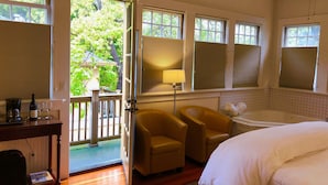 A hotel room with a bed, a desk with a lamp, a chair, a bathtub, and large windows with blinds. - The Inn On First (Napa)