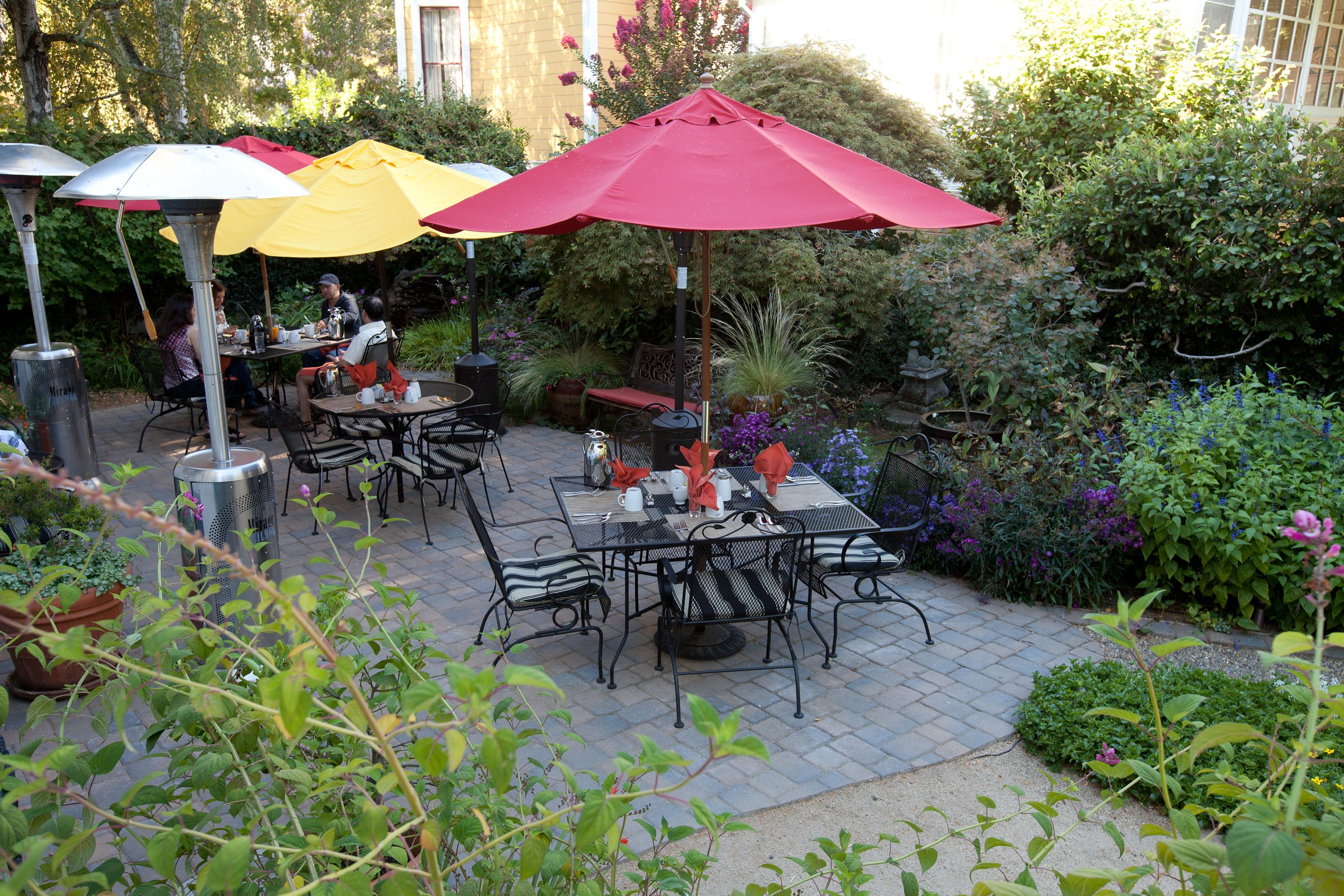 Un patio al aire libre con mesas y sillas, rodeado de plantas en macetas y sombrillas.
