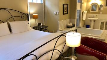 Habitación de hotel con cama, escritorio con lámpara, una silla roja, baño con bañera y espejo.