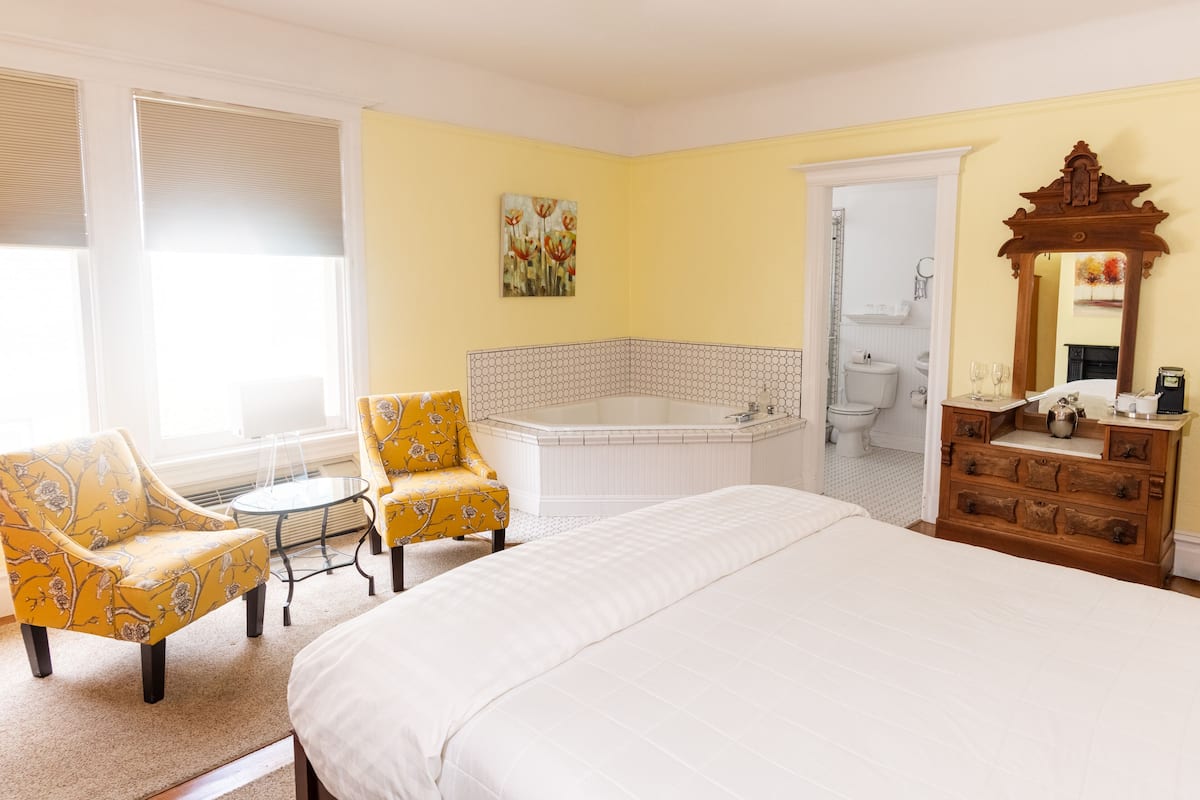 A hotel room with a large bed, two yellow floral armchairs, a small round table, a white bathtub, a wooden dresser, and a mirror.