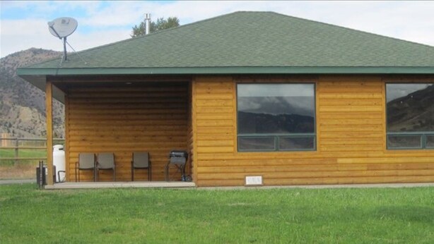A log cabin with a green roof, large windows, and a covered porch area with chairs.