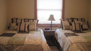 A room with two beds, each with patterned bedding and multiple pillows. There is a nightstand with a lamp and a digital clock between the beds.