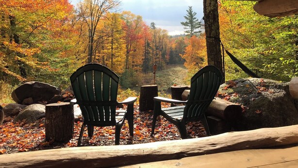 Une cabane en bois avec deux fauteuils Adirondack donnant sur une zone boisée aux couleurs automnales.