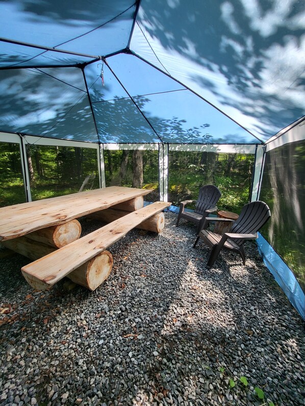 Un espacio de estar al aire libre cubierto con una mesa y sillas de madera, rodeado de grava y árboles.