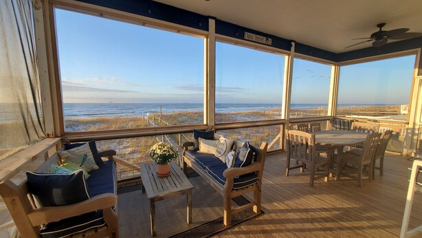A wooden deck with a table and chairs, a cushioned bench, and a view of the beach and ocean.
