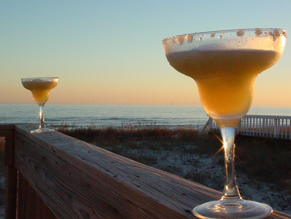 Two cocktail glasses on a wooden deck overlooking a beach at sunset.