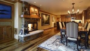 A spacious living room with a stone fireplace, wooden flooring, a dining area with chairs, and a kitchen with wooden cabinets and a chandelier. - Pet Friendly & Luxurious 3 bedroom Aspen Condo (Aspen)