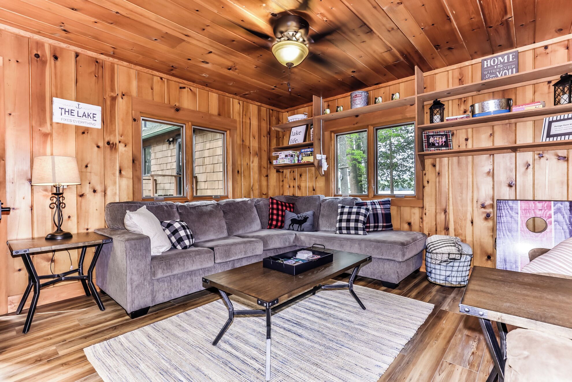 A cozy cabin interior with a gray sectional sofa, a wooden coffee table, and a rustic ceiling fan.
