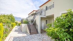 Un edificio de dos pisos con un patio empedrado de piedra, una escalera con barandillas negras y vistas a montañas al fondo.