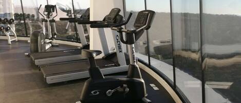 Un gimnasio con cintas de correr y bicicletas estáticas, amplias ventanas con vista a un paisaje urbano.