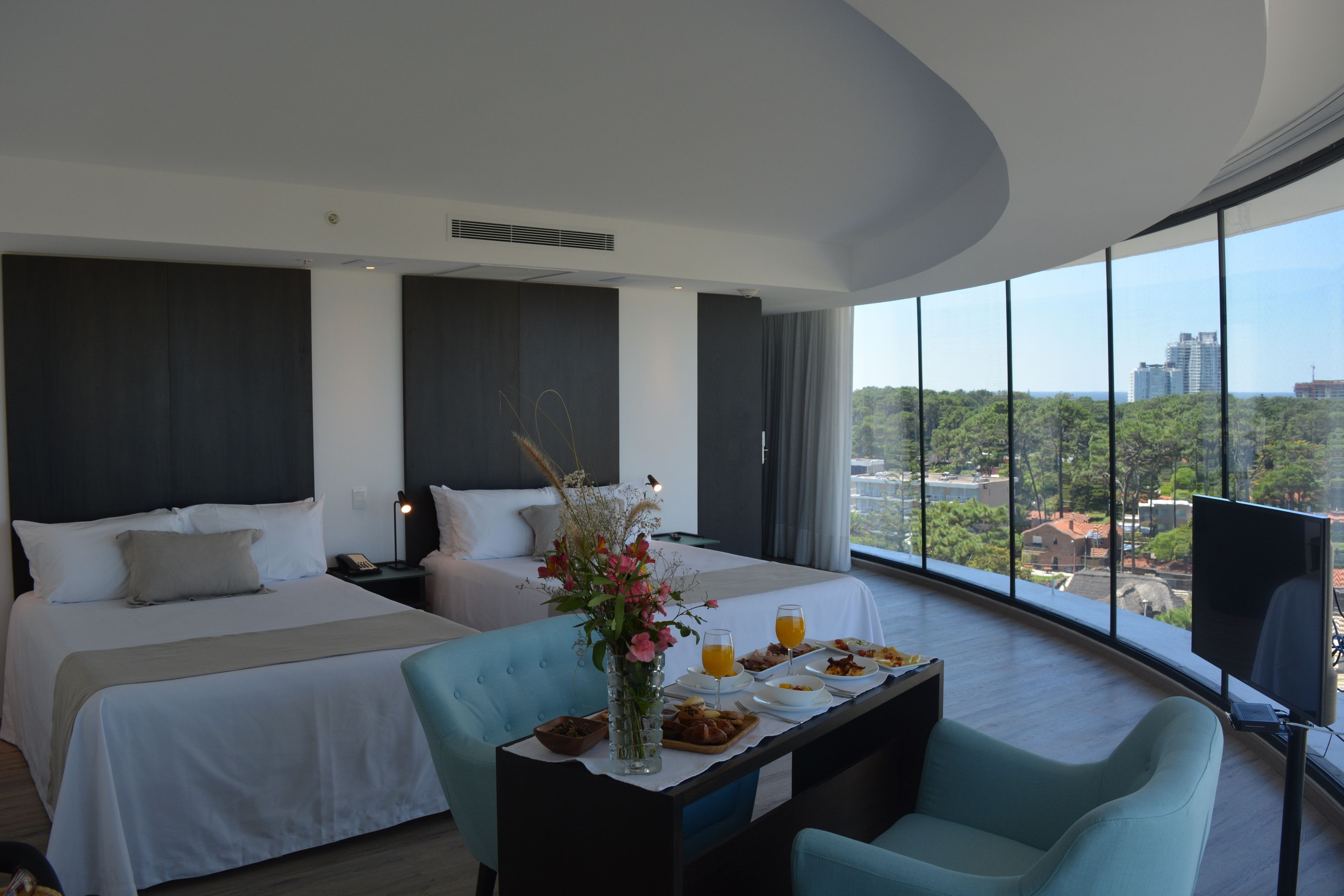 Habitación de hotel con dos camas, una mesa de comedor con desayuno, televisor y un amplio ventanal con vistas a la ciudad.