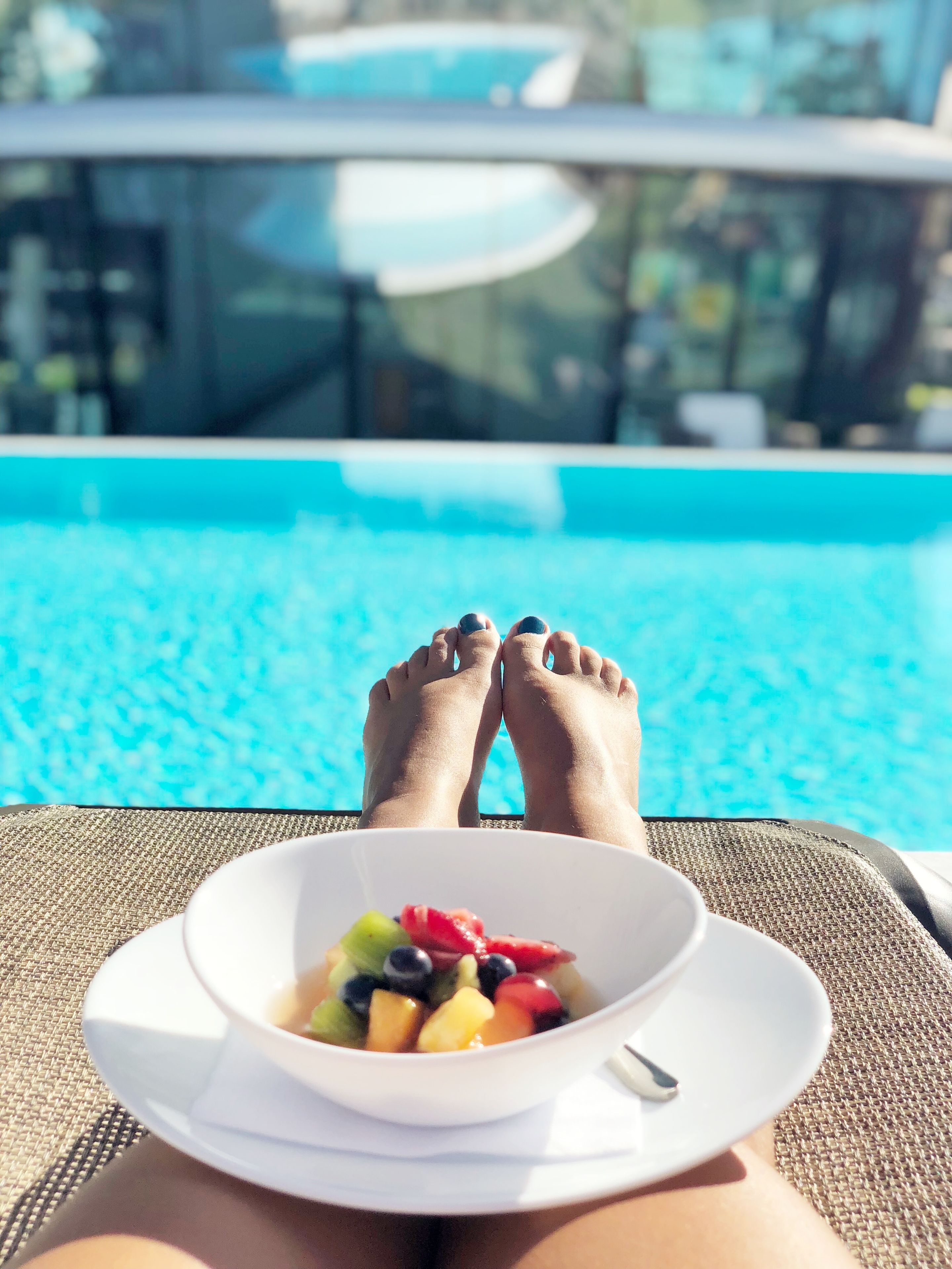 A poolside view with a bowl of fruit and a white plate on a woven chair.