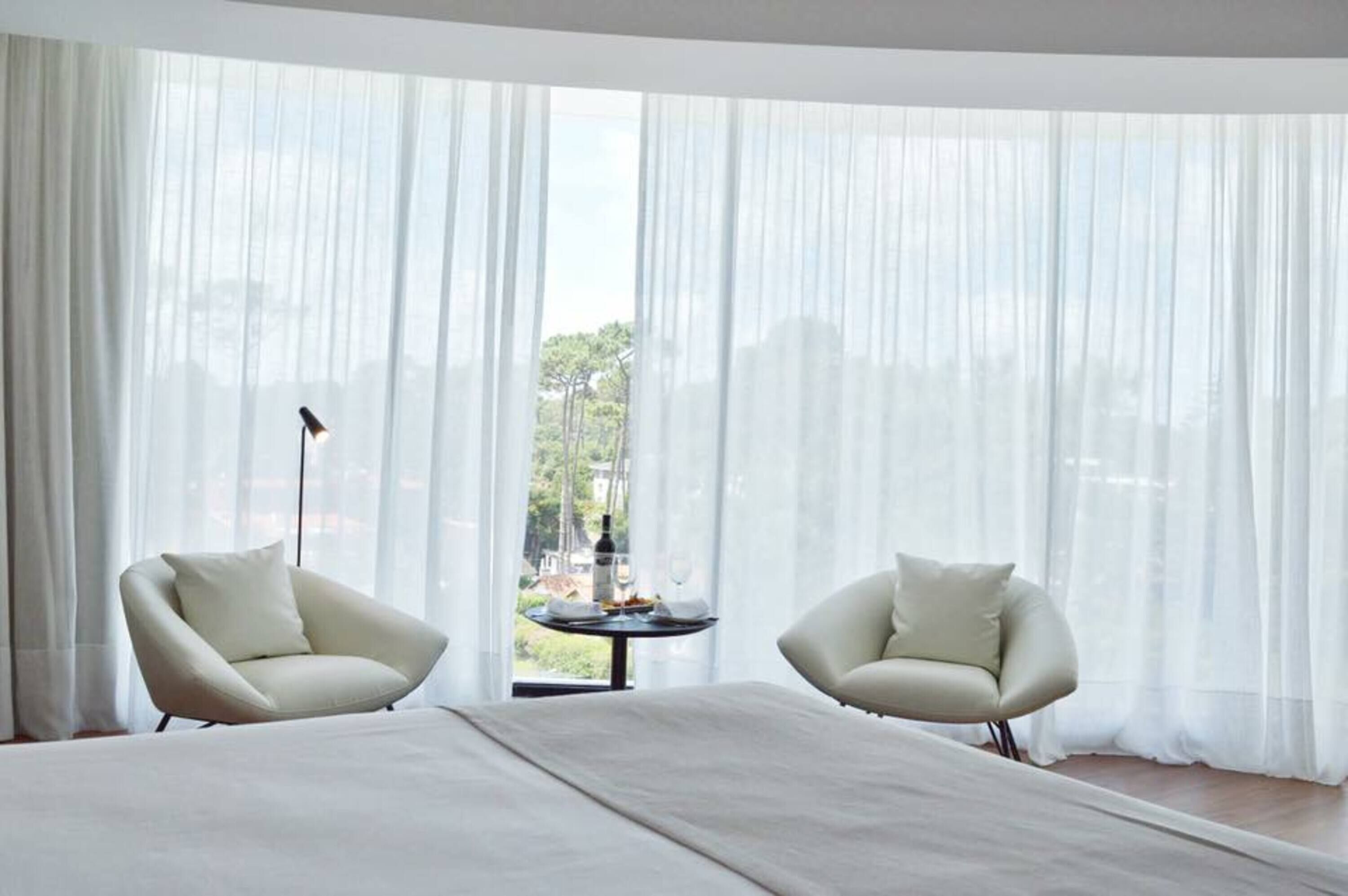 Un dormitorio con una cama, dos sillas, una mesa con una botella y vasos, y cortinas traslúcidas.